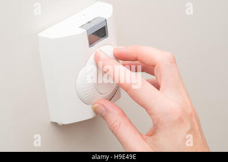 A hand spins the dial on a white wall mounted thermostat on a light coloured wall Stock Photo