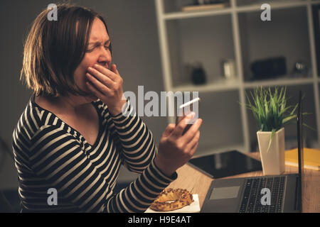 Tired businesswoman working overtime and yawning in office, female person answering to a text message on mobile phone Stock Photo