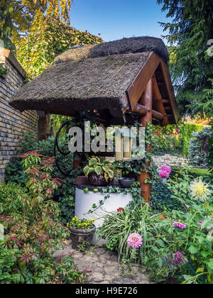 Old rustic thatched well in the garden surrounded by flowers and trees Stock Photo