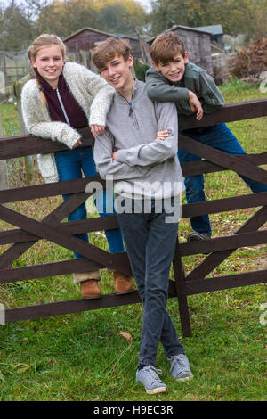 Three children two boys and girl, family brothers and sisters, leaning on a gate in a country garden Stock Photo