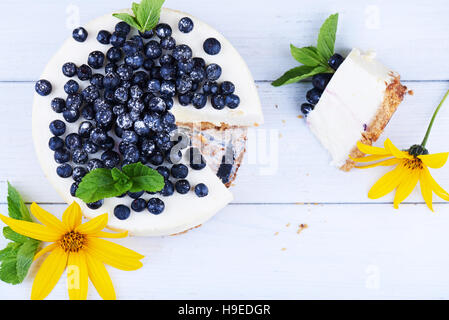 Sweet creamy blueberry cheesecake with fresh blueberries, mint leaves, and yellow flower with a cut piece on a white wooden background with copy space Stock Photo