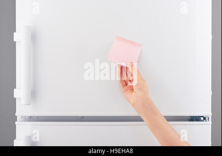Female hand holding blank light pink sticky paper note on white refrigerator door Stock Photo