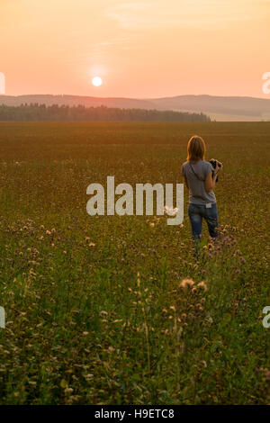 Caucasian woman walking in field at sunset with camera Stock Photo