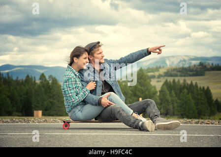 The guy with girl sit on a longboard. Man shows his hand forward. Road in the mountains. Stock Photo