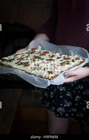 Woman wearing black with painted nails carrying and holding tray bake cake decorated with pecans, rose petals and pumpkin seeds. Stock Photo