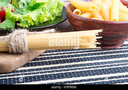 Detail of table with food closeup - lettuce in a black plate, pasta in a wooden bowl and a bunch of spaghetti on a wooden board Stock Photo