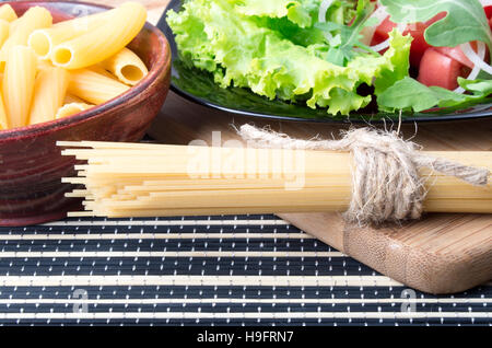 Detail of table with healthy food closeup - lettuce in a black plate, pasta in a wooden bowl and a bunch of spaghetti on a wooden board Stock Photo