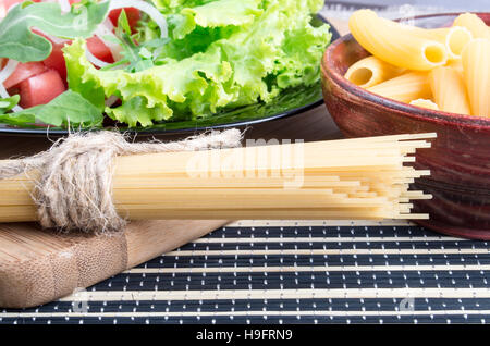 Detail of table with natural food closeup - fresh green salad, pasta in a wooden bowl and a bunch of spaghetti on a wooden board Stock Photo