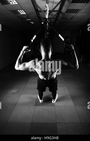 One young handsome brave curly bearded muscled man with a beard makes Total Resistance eXercises fitness trx straps working out in gym Stock Photo