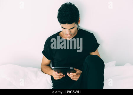 Handsome brazilian guy with cross-earring in black t-shirt and tights is sitting on bed and using digital tablet in the morning after sleeping, white Stock Photo