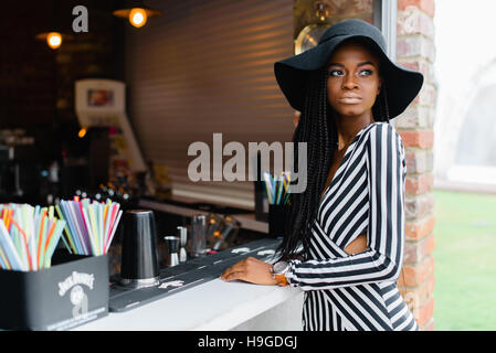 Beautiful african woman in striped dress and black hat is staying in bar. Many straws for cocktails on the bar. Stock Photo