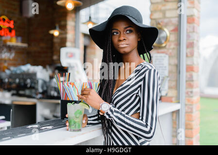 Attractive black girl in bar with cocktail in her hands. Many straws for cocktails on the bar. Ice cocktails. Mohito. Stock Photo
