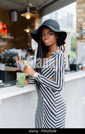 Adorable black girl in bar with cocktail in her hands. Many straws for cocktails on the bar. Ice cocktails. Mohito. Stock Photo