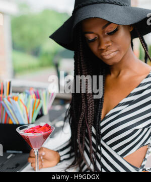 Adorable, charmed black girl in bar with red cocktail in her hands. Many straws for cocktails on the bar. Ice cocktails. Margarita. Stock Photo