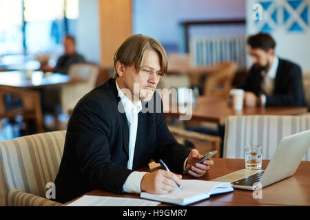 Serious businessman with smartphone making notes in notebook and looking at laptop display Stock Photo