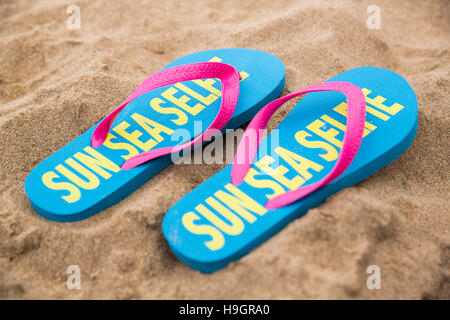 Pair of flip flops with 'Sun Sea Selfie' written on them on a sandy beach. Stock Photo