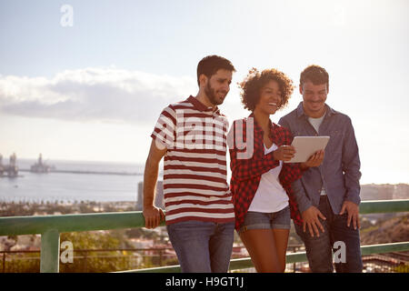 Three laughing good looking friends leaning against a railing looking at a tablet, the girl between the young men is holding Stock Photo