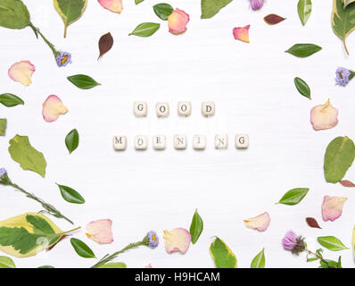 The phrase 'good morning' on cubes in a frame of flowers, petals and green leaves on a white background.Inspirational image.Type flat, top view Stock Photo