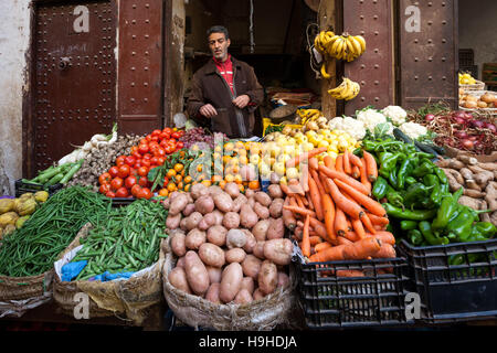 A vendor stands behind his colourful market stall in the medina of Fès, Morocco, bunches of bananas hanging within reach above him. Tomatoes, potatoes, carrots and green peppers are piled in crates and sacks in front of the heavy iron door. Stock Photo