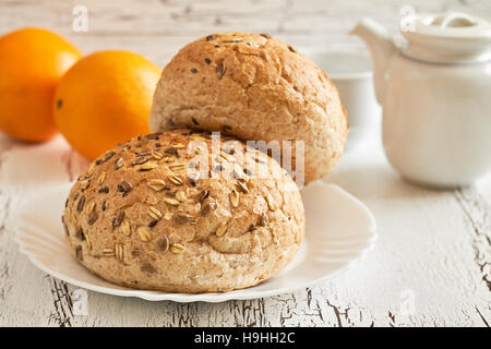 Whole wheat buns with mixed seeds with teapot and oranges in background on white rustic table. Concept image for healthy breakfast Stock Photo