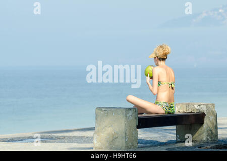 A woman in bikini sitting on a bench overlooking the calm sea drinking a green coconut coco gelado at Arpoador in Rio de Janeiro Stock Photo