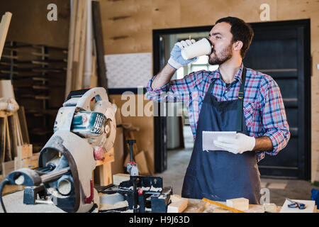 Carpenter taking a coffee break holding notebook in front of circular saw at his workshop Stock Photo