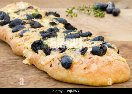 Traditional Italian Stone Baked Focaccia Bread topped with Black Olives, Cheese and Herbs and served on a wooden chopping board Stock Photo