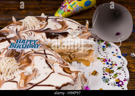 Cut birthday cake with different chocolate ornaments stuffed with condensed milk and nuts on white napkin with colorful confetti and holiday caps on b Stock Photo