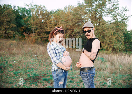 young guy in a cap solar oskah, with beard mustache, and tattooed,  his pregnant girl in dreadlocks, outdoors the fall posing at the camera, Stock Photo