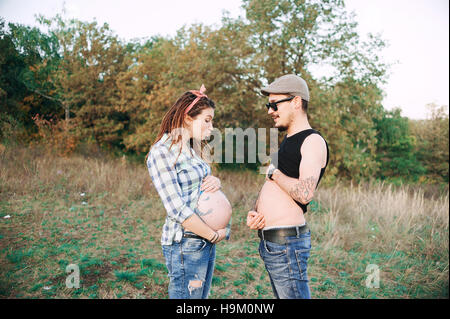 young guy in a cap solar oskah, with beard mustache, and tattooed, his pregnant girl in dreadlocks, outdoors the fall posing at the camera, Stock Photo