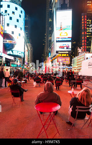 A group of people sitting on chairs on the sidewalk with their backs to the camera watching the big screens at Times Square, New York City, at night. Stock Photo