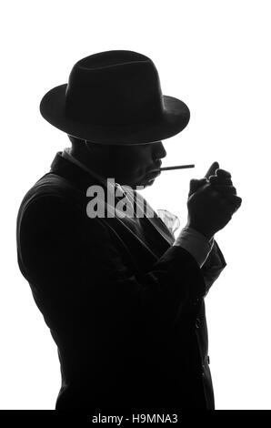 Silhouette of private detective lights cigarette. Agent looks like Al Capone stay side to camera. Police criminal scene in black and white. Gangster studio shot Stock Photo