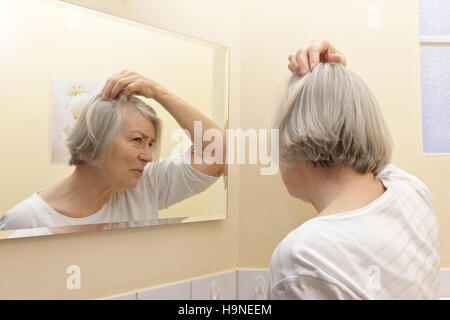 Old woman with thin gray hair and a worried look on her face  examining her beginning baldness  in the mirror of her bathroom Stock Photo