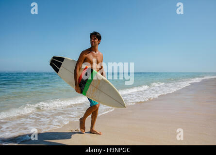 Full length shot of young man with surf board on beach. Handsome caucasian male holding surfboard walking on the sea shore and looking away. Stock Photo