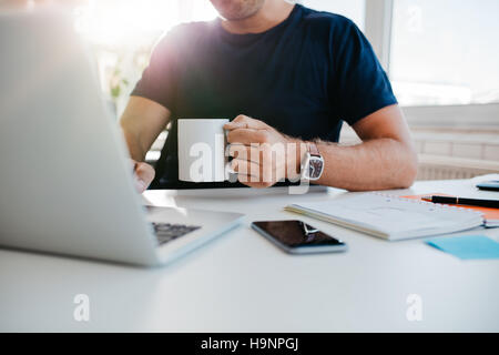 Cropped shot of young man with cup of coffee working on laptop. Business man working at his desk with coffee. Focus on hand. Stock Photo