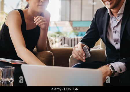 Shot of two young businesspeople sitting together working on laptop. Executives meeting in a office lobby. Man holding mobile phone and woman pointing Stock Photo