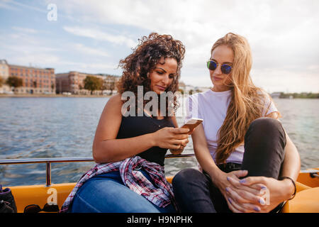 Shot of two young women sitting on boat and looking at mobile phone. Female friends using smart phone by the lake. Stock Photo