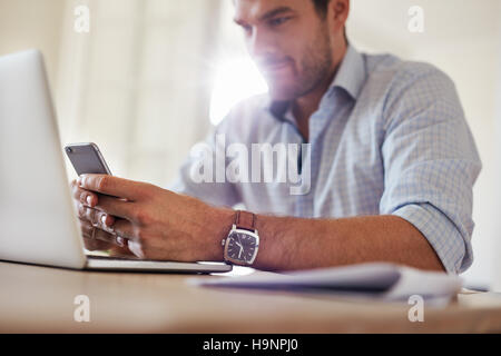 Shot of young businessman using mobile phone at home. Man sitting at table with laptop and reading text message on his smart phone. Stock Photo