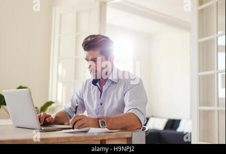 Indoor shot of young man working on laptop in the office and writing down notes. Business man working from home. Stock Photo