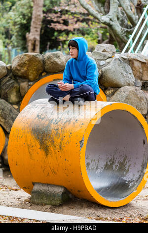 Japan, Akashi. Kazki, early teenage caucasian boy in blue hoodie sitting cross-legged on top of yellow tunnel section in play-park. Alone. Thoughtful. Stock Photo