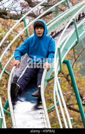 Japan, Akashi. Kazki, early teenage caucasian boy, 13-14 years old, in blue hoodie sliding on curved slide towards viewer. Eye-contact. Stock Photo