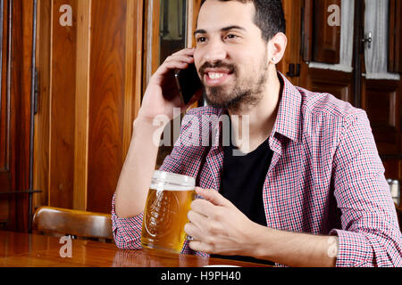 Portrait of young latin man talking on the phone with glass of beer and snacks in a bar. Indoors. Stock Photo