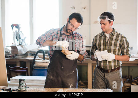 Happy male carpenter showing something to coworker at his notebook papers in workshop Stock Photo