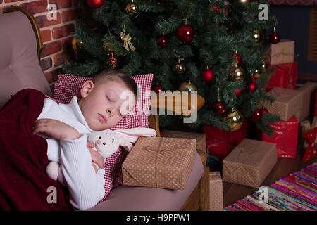 Boy sleeping under a blanket on the sofa near the Christmas tree with gifts Stock Photo