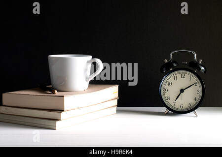 Retro alarm clock beside a cup of coffee on top the books with pencil on grunge black background on white table. Stock Photo