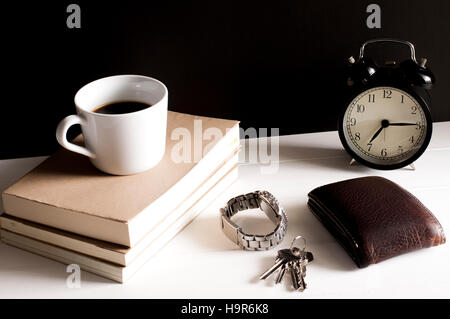 Wallet,watch and key chain beside a cup of coffee on top the books and retro alarm clock on white table in front of grunge black background. Stock Photo