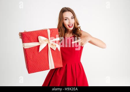 Happy excited woman in red dress pointing finger at big gift box isolated over white background Stock Photo