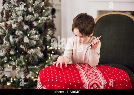 boy calls Santa while sitting on a big green armchair at home over chirstmas tree background Stock Photo