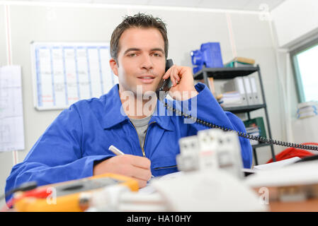 Man in overalls on telephone, sat at desk Stock Photo
