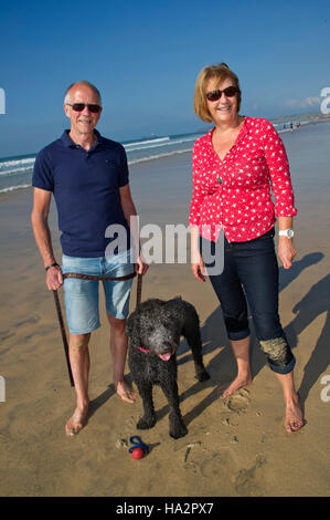 A mature couple (male & female) wearing Summer clothes with their dog having fun on a sunny Cornish beach. Stock Photo